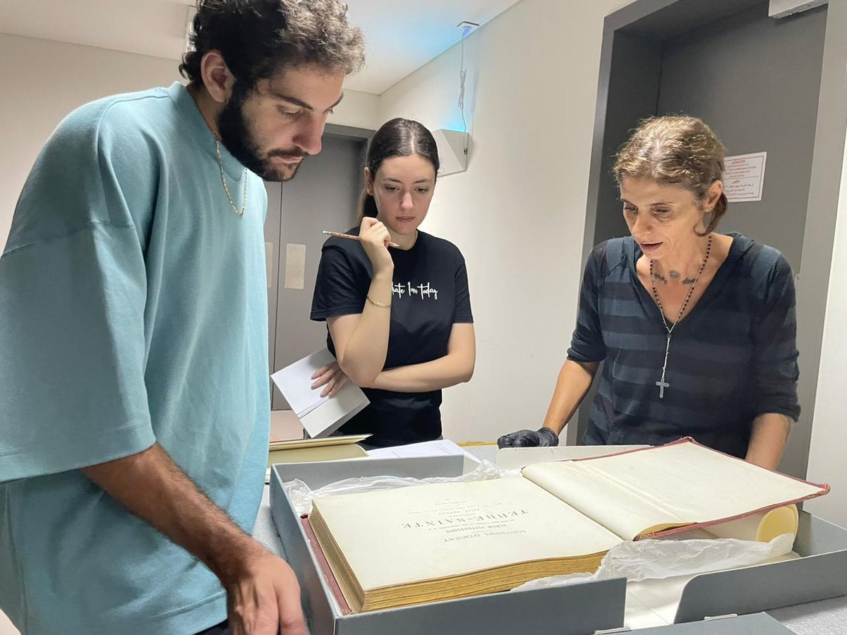 Three people examining an old album, “Souvenirs d’Orient. Album pittoresque … Terre-Sainte”, kept in an archival box