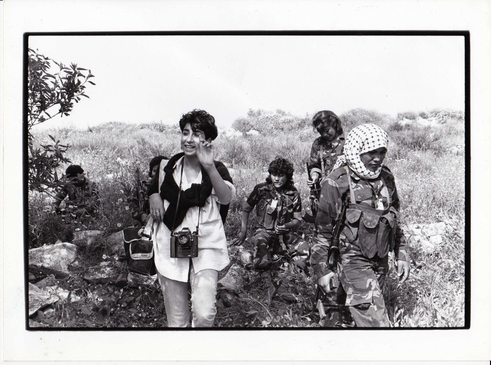 Black-and-white photograph: a photographer, camera around her neck, walking among young armed fighters in a field